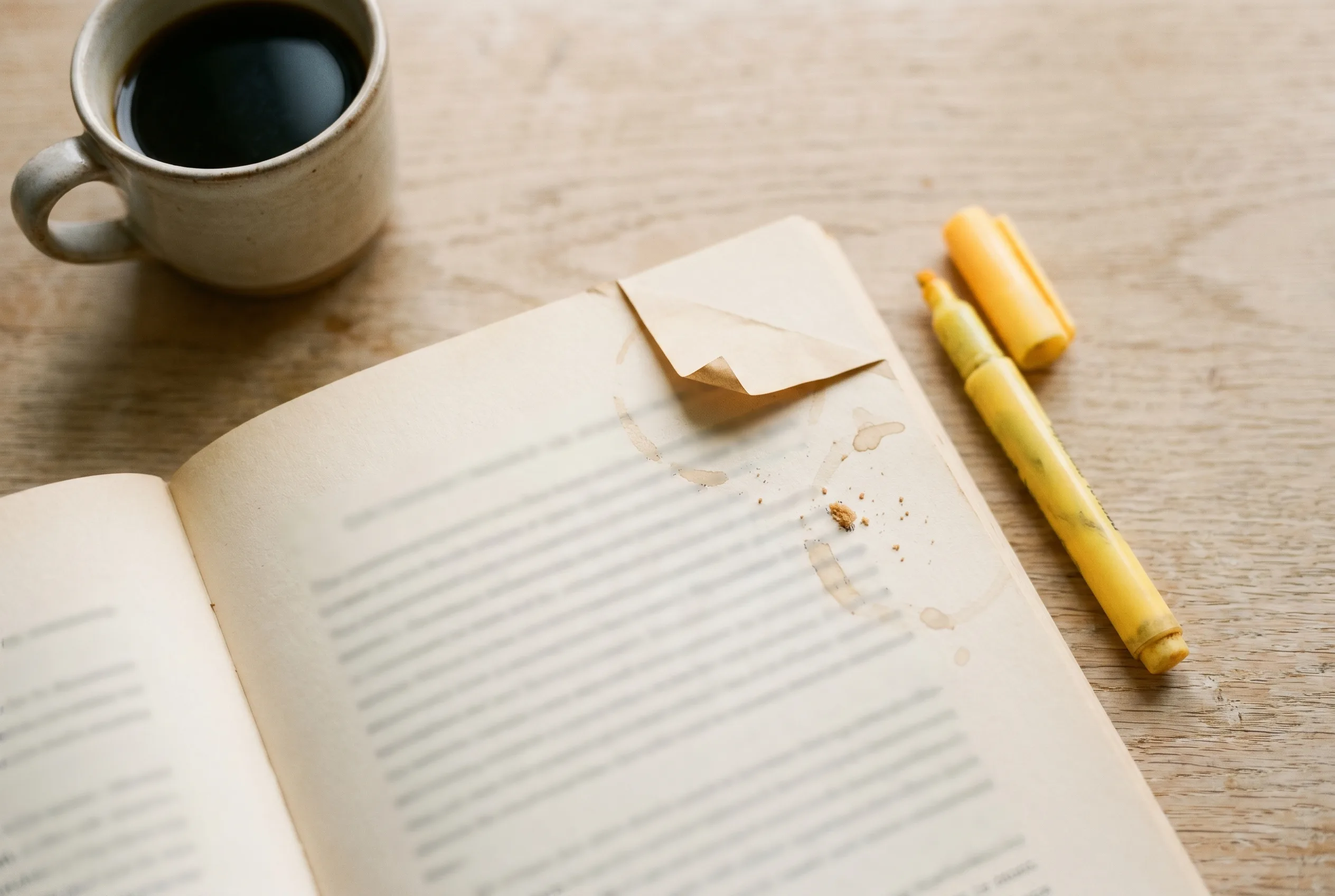 Overhead macro of an open paperback with a yellow highlighter across a printed page, a folded paper bookmark, and a small ceramic mug of black coffee out of focus in the corner.