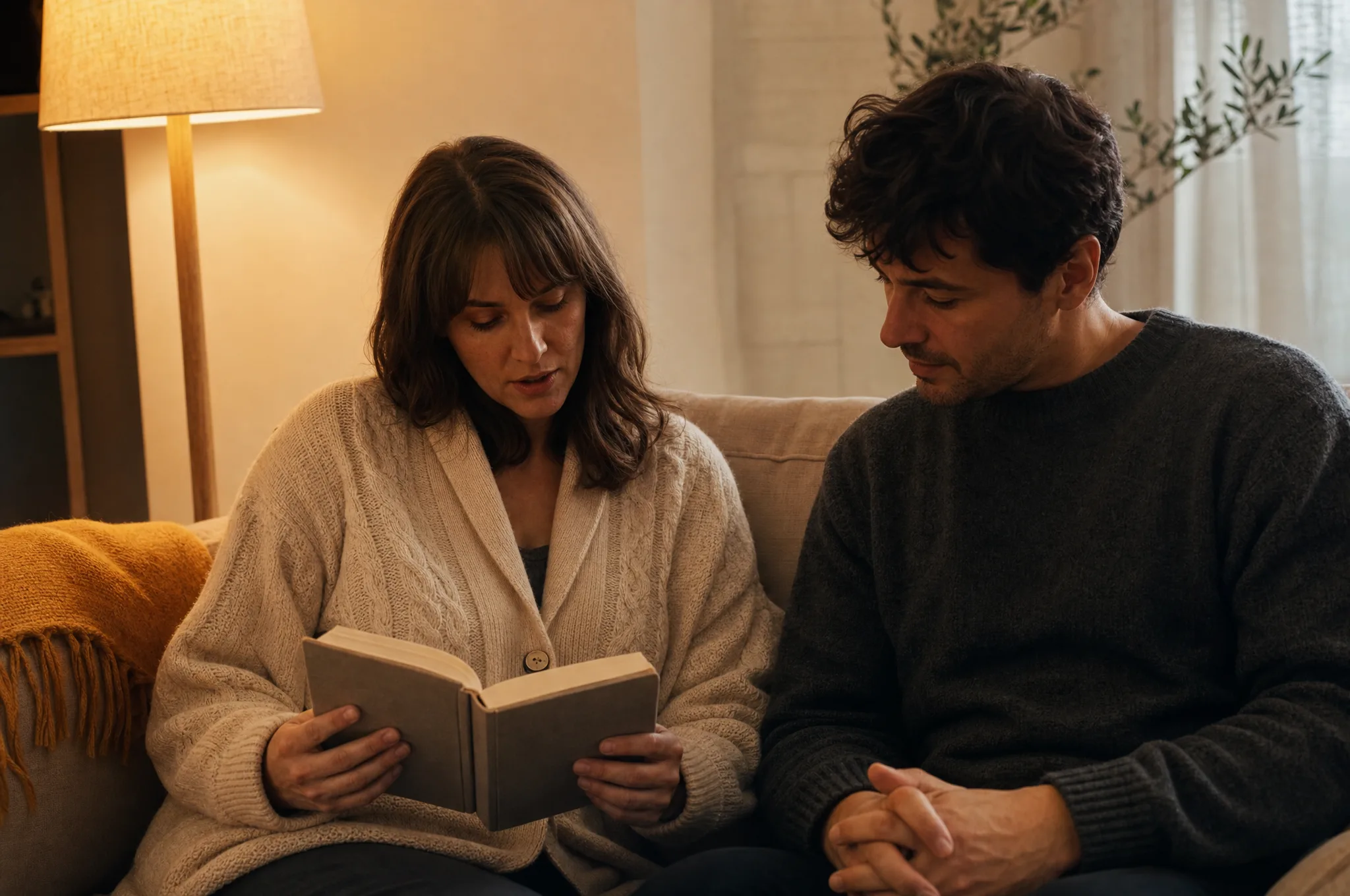 Two people on a sand-colored couch in warm lamp light. A woman reads aloud from a clothbound paperback; a man listens with hands clasped in his lap.